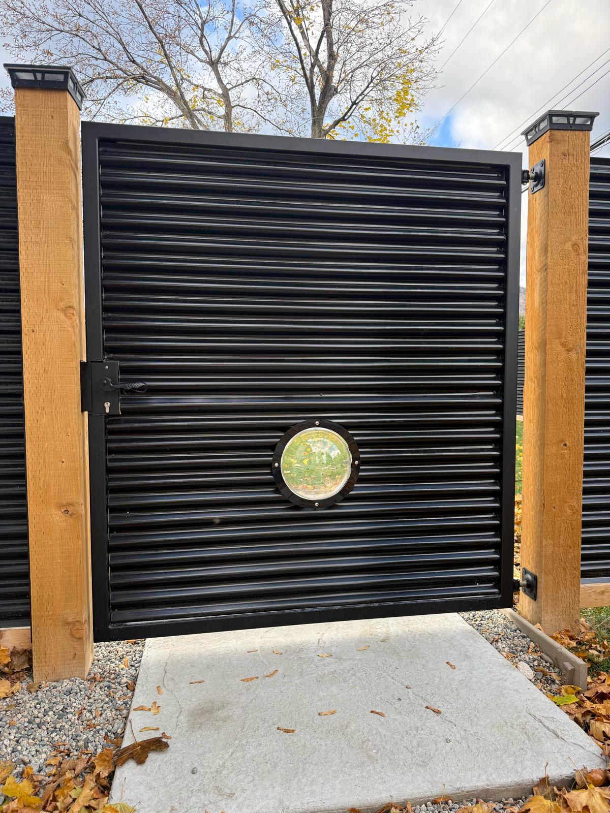Black corrugated metal privacy gate with round pet peek window on 6×6 treated posts.